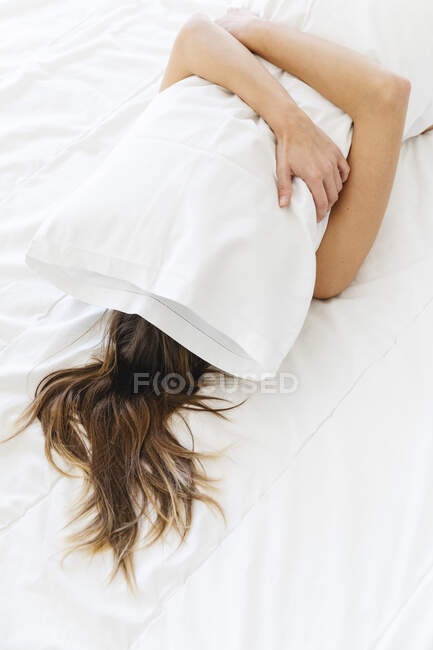 Young woman covering face with white pillow while lying on bed at home — Stock Photo