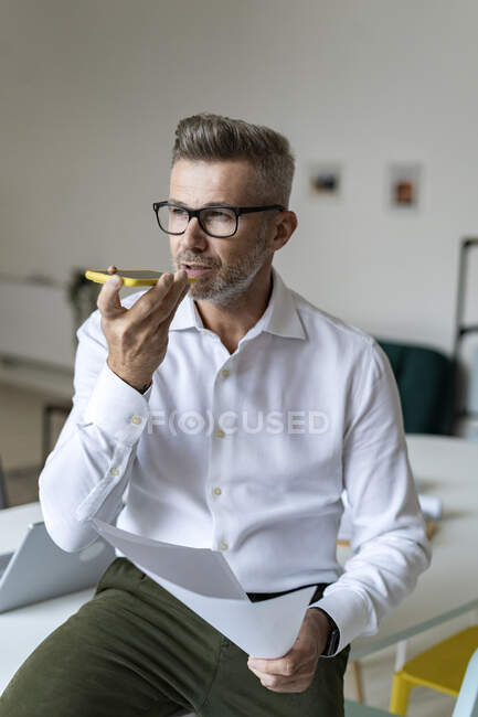 Geschäftsmann hält Dokument in der Hand, während er im Büro mit dem Smartphone spricht — Stockfoto