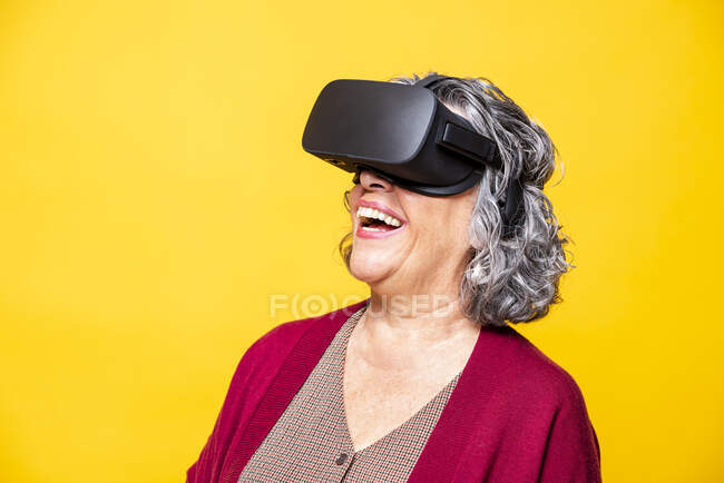 Mujer mayor riendo mientras usa auriculares de realidad virtual sobre fondo amarillo - foto de stock