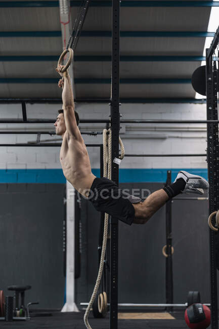 Jeune athlète masculin se balançant tout en tenant des anneaux de gymnastique dans la salle de gym — Photo de stock