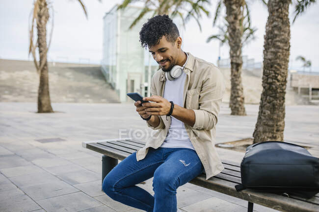 Man surfing net through mobile phone while sitting by backpack on bench — Stock Photo