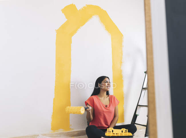 Smiling young woman with paint roller sitting by ladder at home — Stock Photo
