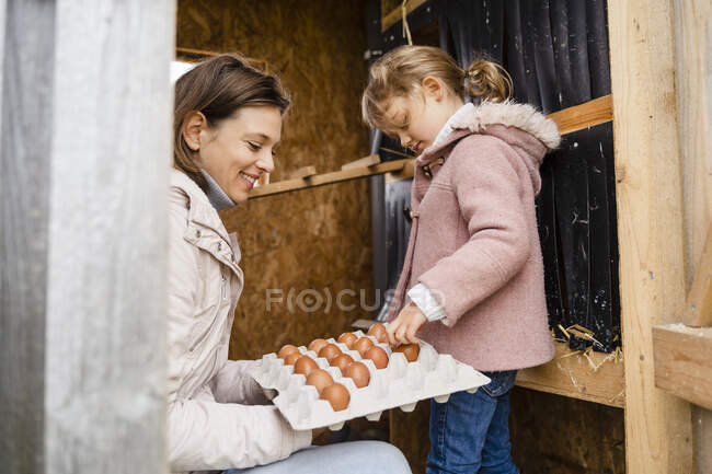 Mãe segurando recipiente enquanto meninas organizando ovos na fazenda — Fotografia de Stock