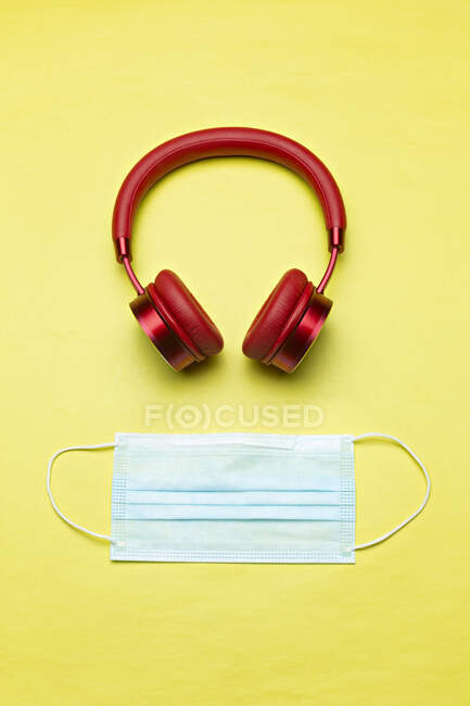 Studio shot of red wireless headphones and protective face mask lying against yellow background — Stock Photo