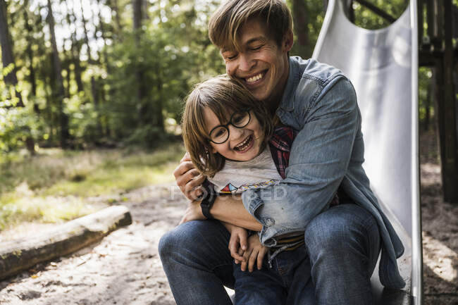 Sonriente padre con los ojos cerrados abrazando hijo en diapositiva en el bosque - foto de stock