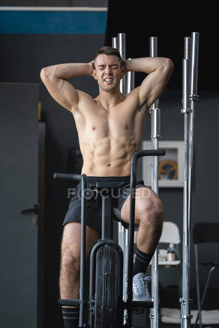 Atleta masculino cansado con las manos detrás de la cabeza sentado en el cross trainer en el gimnasio - foto de stock