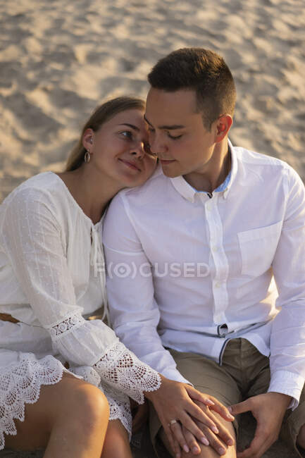 Mujer joven inclinada sobre el hombro del novio mientras está sentada en la arena - foto de stock