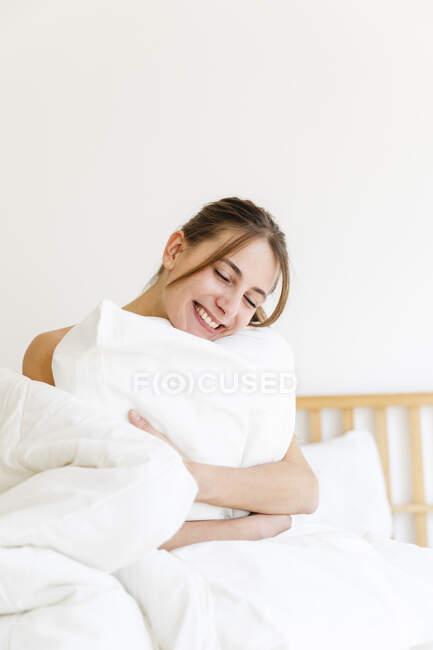 Happy young woman with eyes closed embracing white pillow at home — Stock Photo