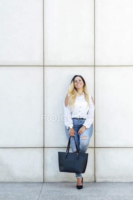 Mujer de negocios sonriente con bolsa apoyada en la pared - foto de stock