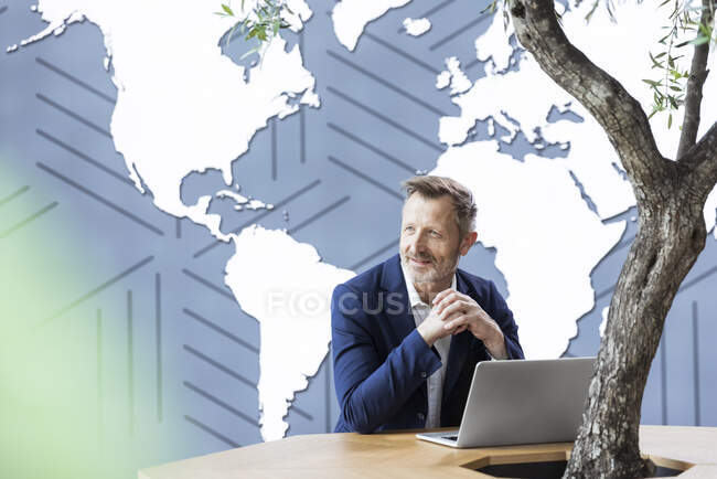 Uomo d'affari sorridente con le mani giunte seduto alla scrivania in ufficio — Foto stock