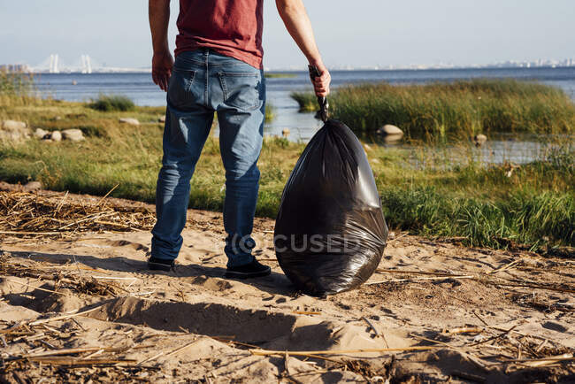 Взрослый волонтер держит мусорный мешок на берегу реки в солнечный день — стоковое фото