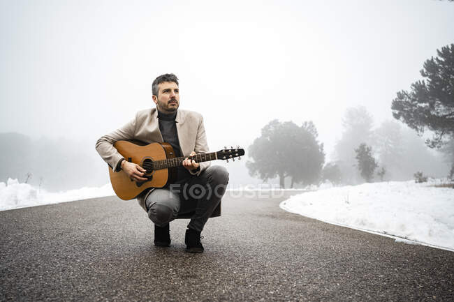 Homem maduro tocando guitarra enquanto se agacha na estrada — Fotografia de Stock