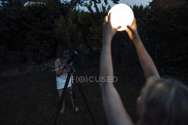 Chica mirando linterna iluminada a través del telescopio durante la puesta del sol - foto de stock
