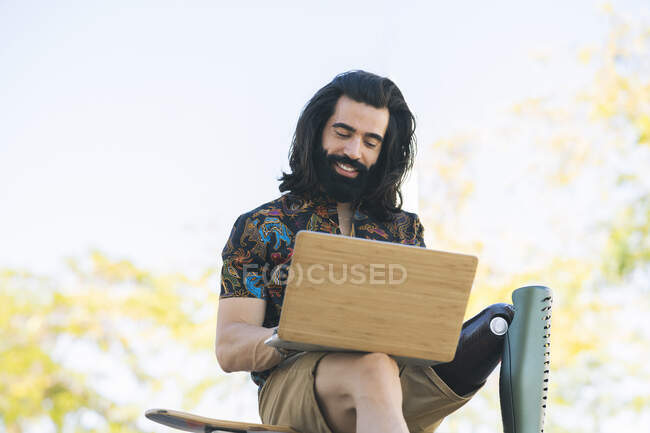 Усміхнений інвалідний чоловік з довгим волоссям використовує ноутбук у громадському парку — стокове фото