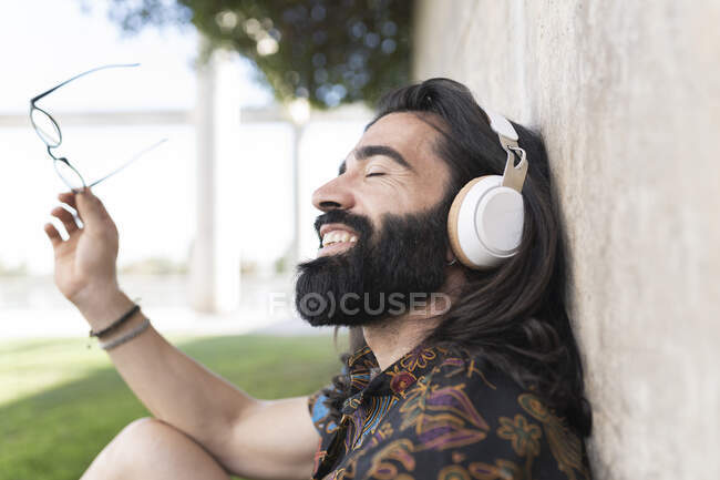 Heureux hipster homme portant des écouteurs tenant des lunettes dans le parc — Photo de stock