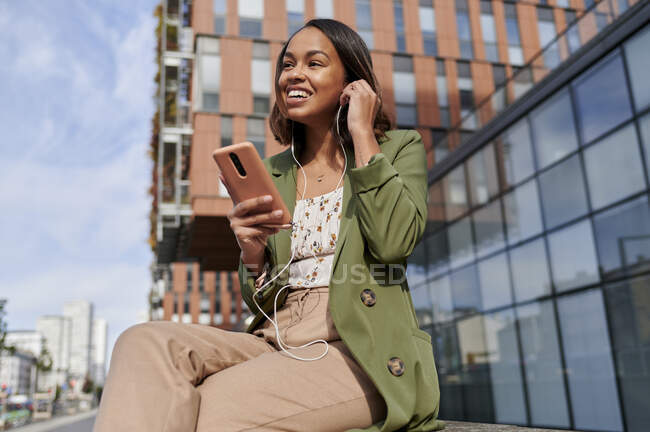 Mulher sorridente com telefone celular usando fones de ouvido na cidade — Fotografia de Stock