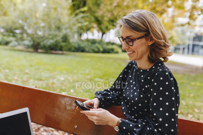 Smiling female freelancer text messaging through smart phone in public park — Stock Photo