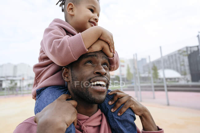 Sonriente padre mirando a su hijo sentado en los hombros en la cancha deportiva - foto de stock