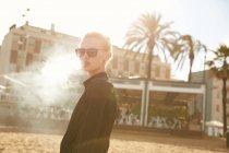 Portrait of woman in sunglasses smoking electronic cigarette on beach in barcelona — Stock Photo
