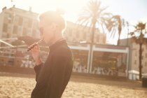 Vista lateral de mujer en gafas de sol fumar cigarrillo electrónico en la playa en barcelona - foto de stock
