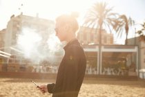 Vue latérale de la femme en lunettes de soleil fumant cigarette électronique sur la plage à Barcelone — Photo de stock