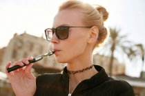 Retrato de mujer joven en gafas de sol fumando cigarrillo electrónico en la calle en barcelona - foto de stock