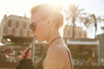 Vista lateral da mulher em óculos de sol fumar cigarro eletrônico na rua em barcelona — Fotografia de Stock