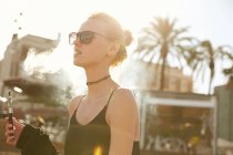 Portrait of young woman in sunglasses smoking electronic cigarette on street in barcelona — Stock Photo