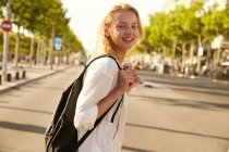 Joven mujer sonriente caminando con el bolso en la calle y mirando a la cámara en barcelona - foto de stock