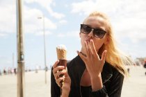 Atractivo turista rubio en gafas de sol lamiendo dedo y sosteniendo cono de helado en la calle - foto de stock