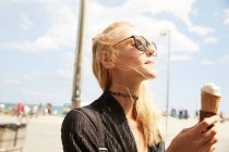 Vue latérale de attrayant touriste blonde dans les lunettes de soleil manger de la crème glacée sur la rue — Photo de stock
