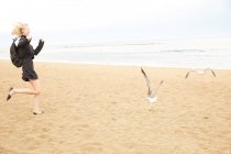 Vista lateral da mulher feliz em vestido preto e saco correndo atrás de gaivotas na praia de areia — Fotografia de Stock