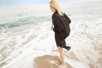 Vue arrière de belle femme en robe noire et sac debout sur la plage de sable de la mer — Photo de stock