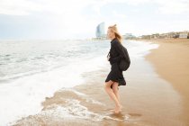 Vista laterale di bella donna in abito nero e borsa in piedi sulla spiaggia sabbiosa del mare — Foto stock