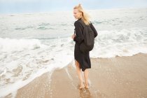 Vista posteriore di bella donna in abito nero e borsa in piedi sulla spiaggia sabbiosa del mare — Foto stock