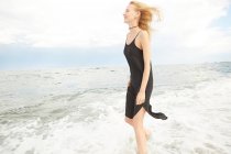 Vista lateral de mulher bonita sorridente em vestido preto em pé no mar — Fotografia de Stock