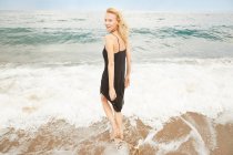 Belle femme blonde en robe noire debout dans la mer — Photo de stock