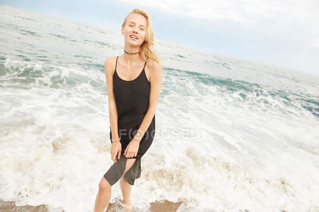 Bela mulher em vestido preto de pé no mar e olhando para a câmera — Fotografia de Stock