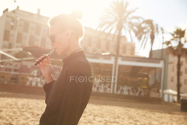 Seitenansicht einer Frau mit Sonnenbrille, die am Strand von Barcelona elektronische Zigarette raucht — Stockfoto