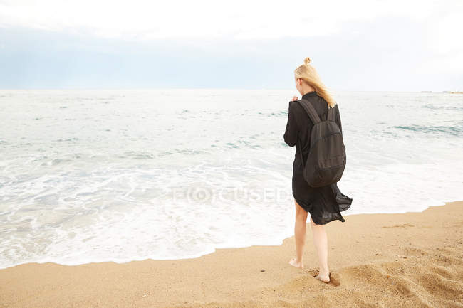 Visão traseira da mulher em vestido preto e saco de pé na areia perto do mar — Fotografia de Stock