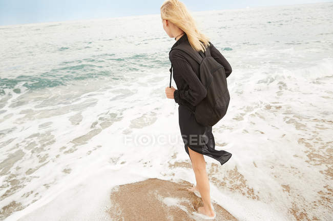 Back view of beautiful woman in black dress and bag standing on sandy sea beach — Stock Photo
