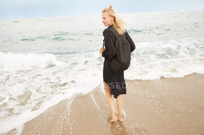 Vue arrière de belle femme en robe noire et sac debout sur la plage de sable fin — Photo de stock