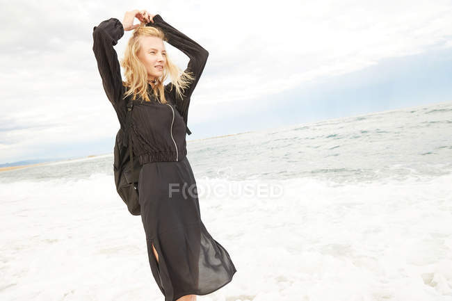 Hermosa mujer en vestido negro y bolsa de pie en el mar y mirando hacia otro lado - foto de stock