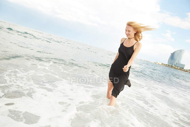 Mujer atractiva sonriente en vestido negro caminando en la playa del mar - foto de stock