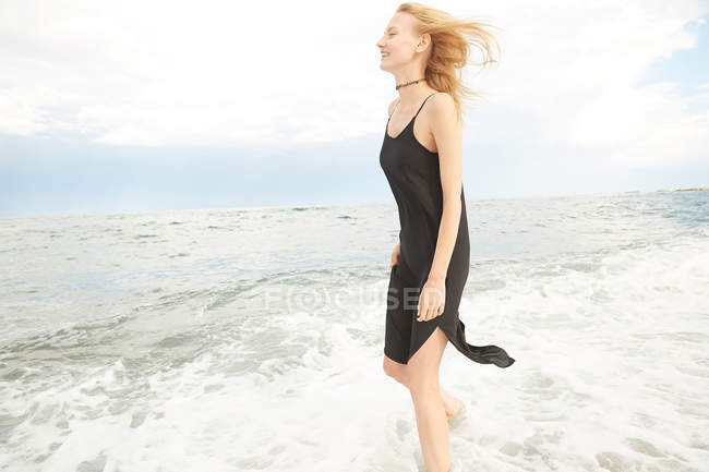 Vista lateral de mulher bonita sorridente em vestido preto em pé no mar — Fotografia de Stock