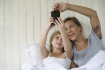 Madre e hija posando para selfie en la cama - foto de stock