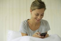 Lächelnde Frau sitzt im Bett und schreibt SMS auf dem Smartphone — Stockfoto