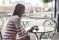 Молода жінка сидить у тротуарному кафе, сміється зі смартфона — стокове фото
