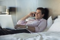Homme couché au lit, utilisant un téléphone portable et un ordinateur portable — Photo de stock