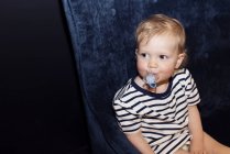 Retrato de niño pequeño sosteniendo chupete en la boca sentado en el sofá - foto de stock